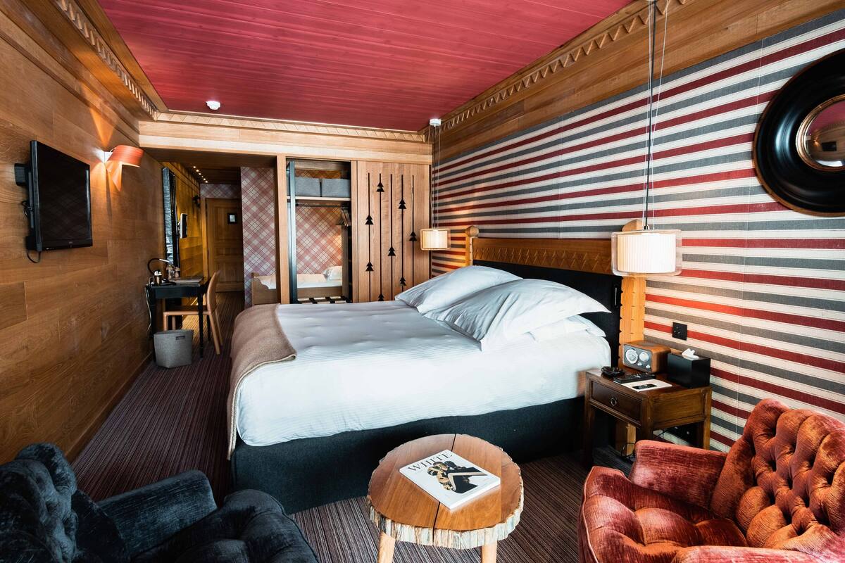 A hotel room with a bed, a wooden headboard, a nightstand, a red armchair, a small wooden stool, and a television mounted on the wall.