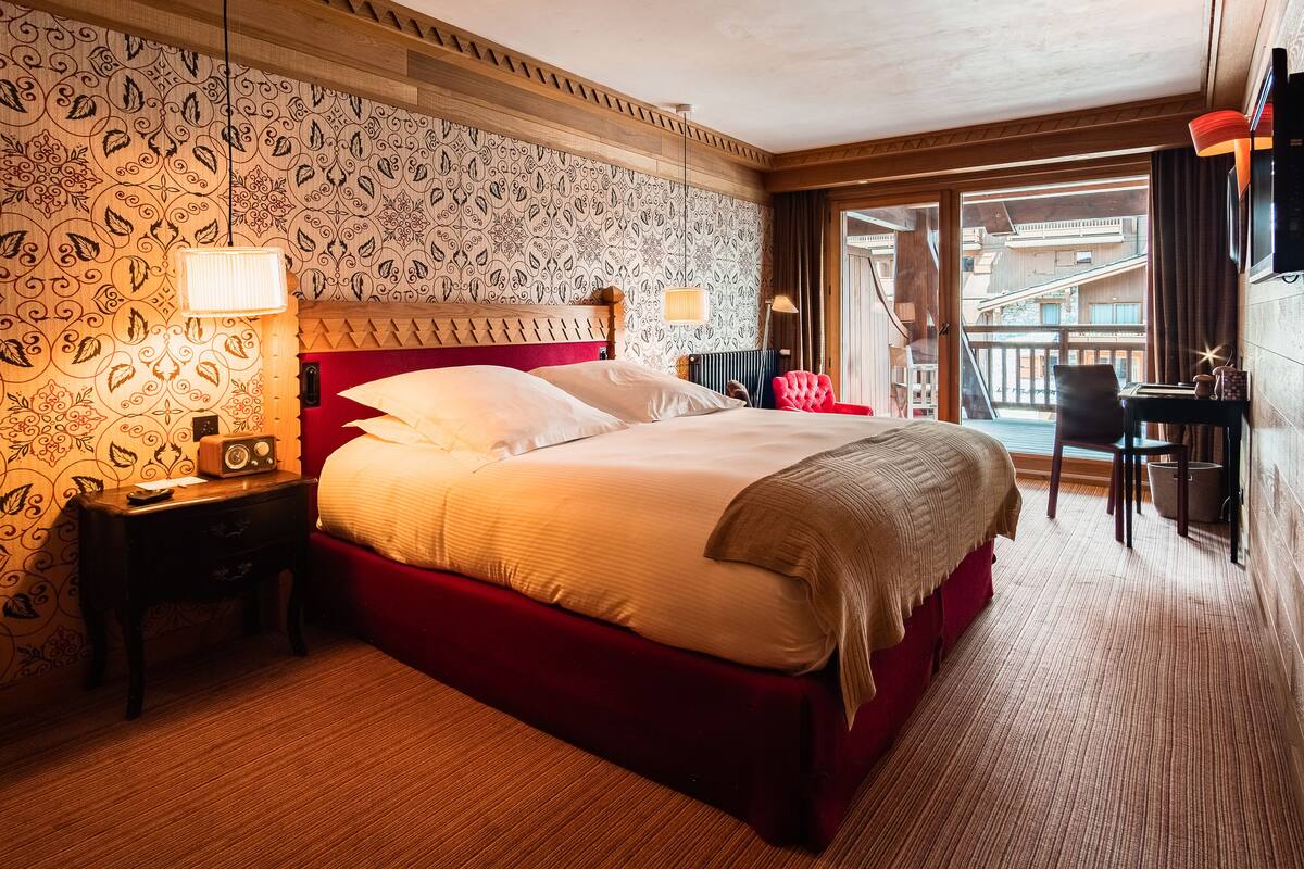 A hotel room with a large bed, a wooden headboard, a red accent wall, a patterned wallpaper, a desk with a chair, and a view of the outside balcony.
