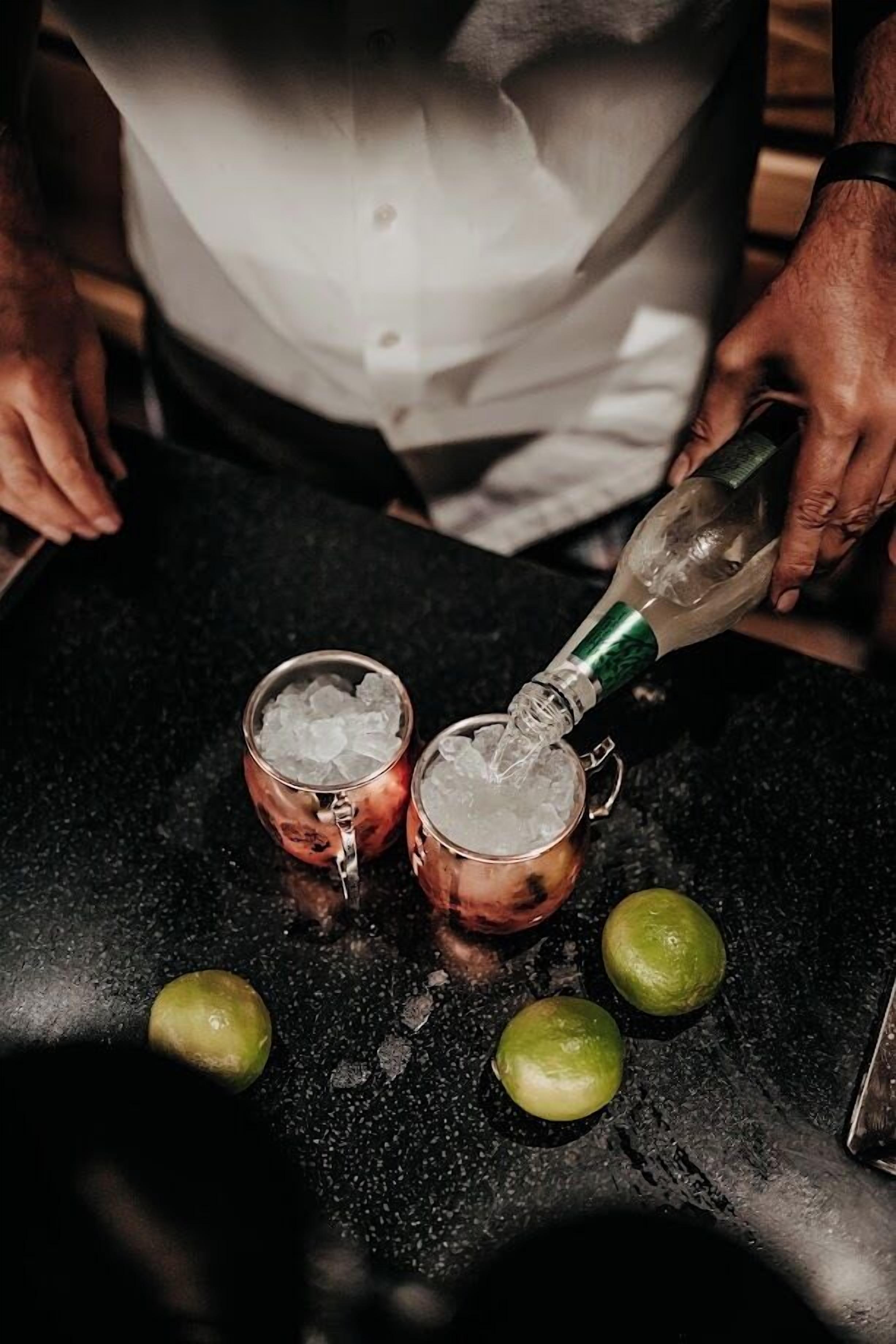 A person pouring a clear liquid into two metal cups filled with ice on a black countertop.