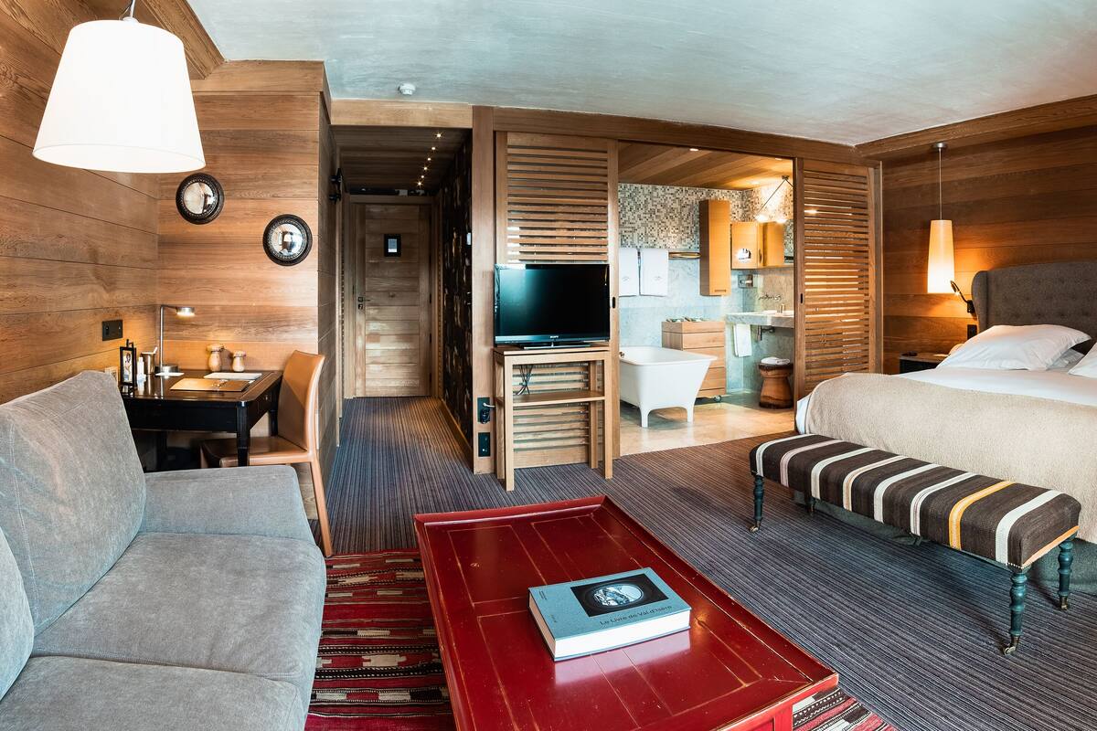 A hotel room with a sofa, a red coffee table with a book, a bed with striped bedding, a TV, and wooden walls.