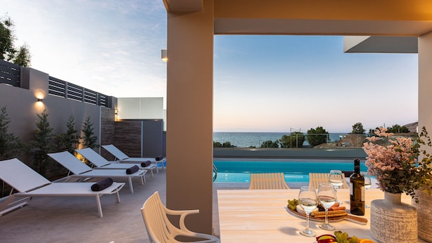 A poolside area with lounge chairs, a table with fruit and wine, and a view of the sea. - Lagoon Seaside Villa, Walking distance to the beach & shops!Car Free holiday! (Panormos in Rethymno)