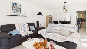 Una sala de estar moderna con dos sofás, una mesa de centro con cruasanes y bebidas, y una zona de comedor al fondo.
