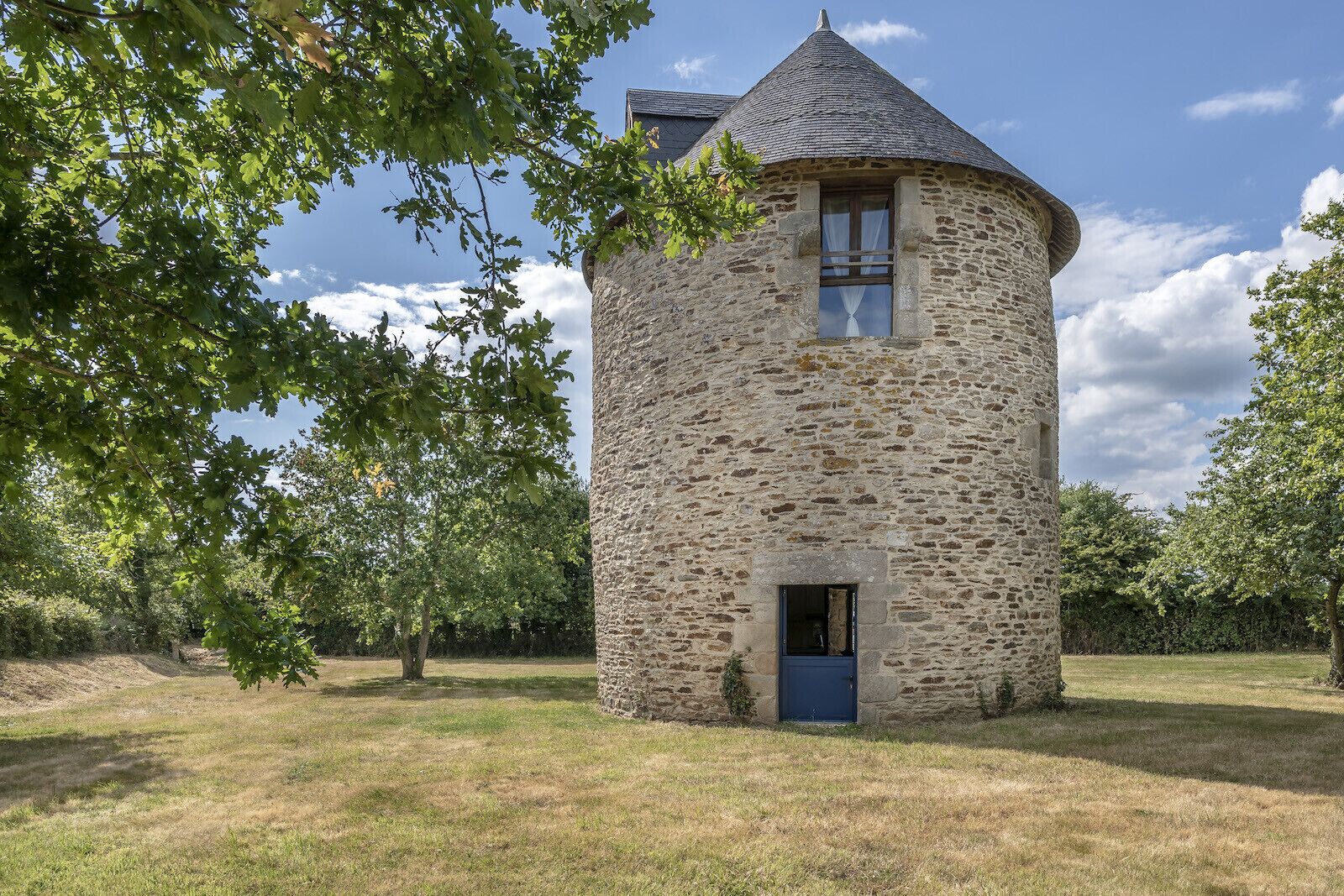Ein steinerner Turm mit einem spitz zulaufenden Dach, einem kleinen Fenster und einer blauen Tür.