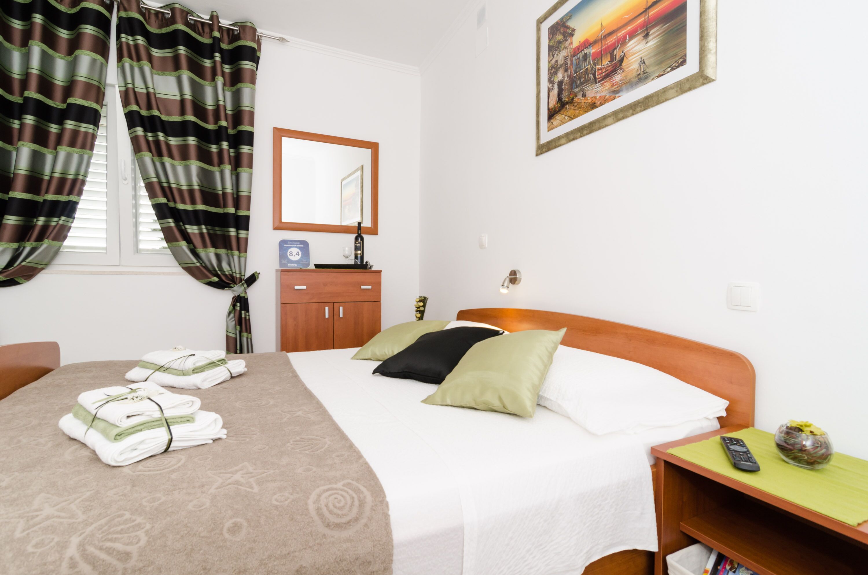 A hotel room with a bed, a wooden nightstand, a mirror, a window with curtains, and a painting on the wall.