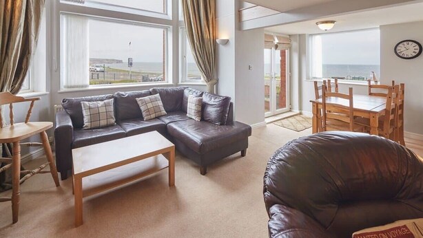 A modern living room with a sectional sofa, a wooden coffee table, and a leather armchair. There are large windows with curtains, a dining area with wooden chairs, and a view of the sea. - Host & Stay | Sunset View (Whitby)