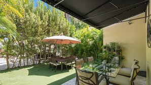 An outdoor patio with a glass table, chairs, and a large umbrella. There are potted plants and a lawn. - Villa Venezia BB (Miami Beach)
