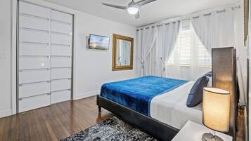Un dormitorio con cama, ventilador de techo, mesita de noche, lámpara, ventana con cortinas, espejo y armario.
