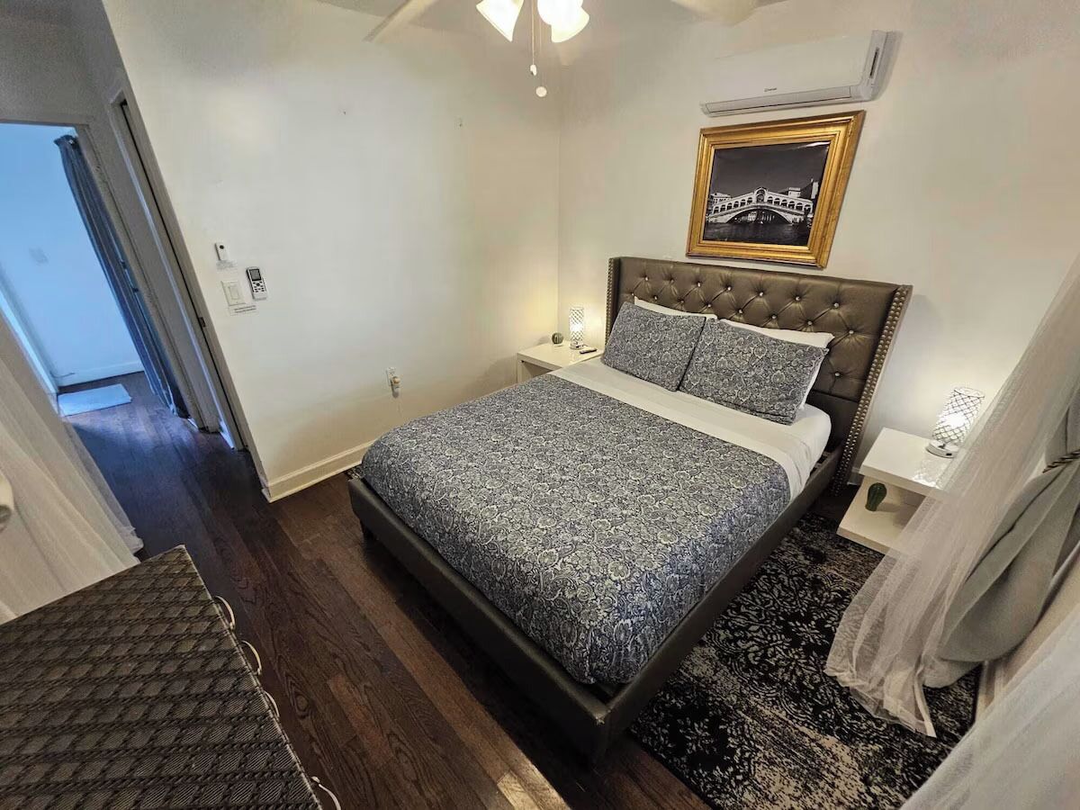 A bedroom with a bed, bedside tables, a nightstand, a wall-mounted air conditioner, and a framed picture.