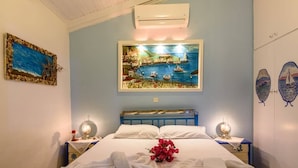 A bedroom with a bed, a painting of a coastal scene, a nightstand with a lamp, and a wall-mounted air conditioner. - Blue Seaside Retreat - Fully Equipped Garden Gem (Chrani)