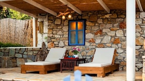 A stone-walled outdoor seating area with two wooden lounge chairs and a small table. - Blue Seaside Retreat - Fully Equipped Garden Gem (Chrani)