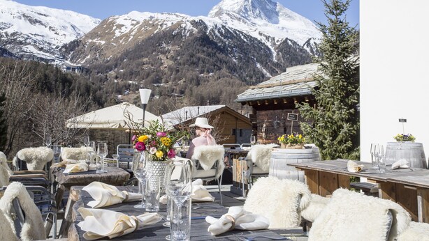 An outdoor dining area with tables set for two, chairs with fur throws, and a mountain backdrop. - Chalet Eggen (Zermatt)