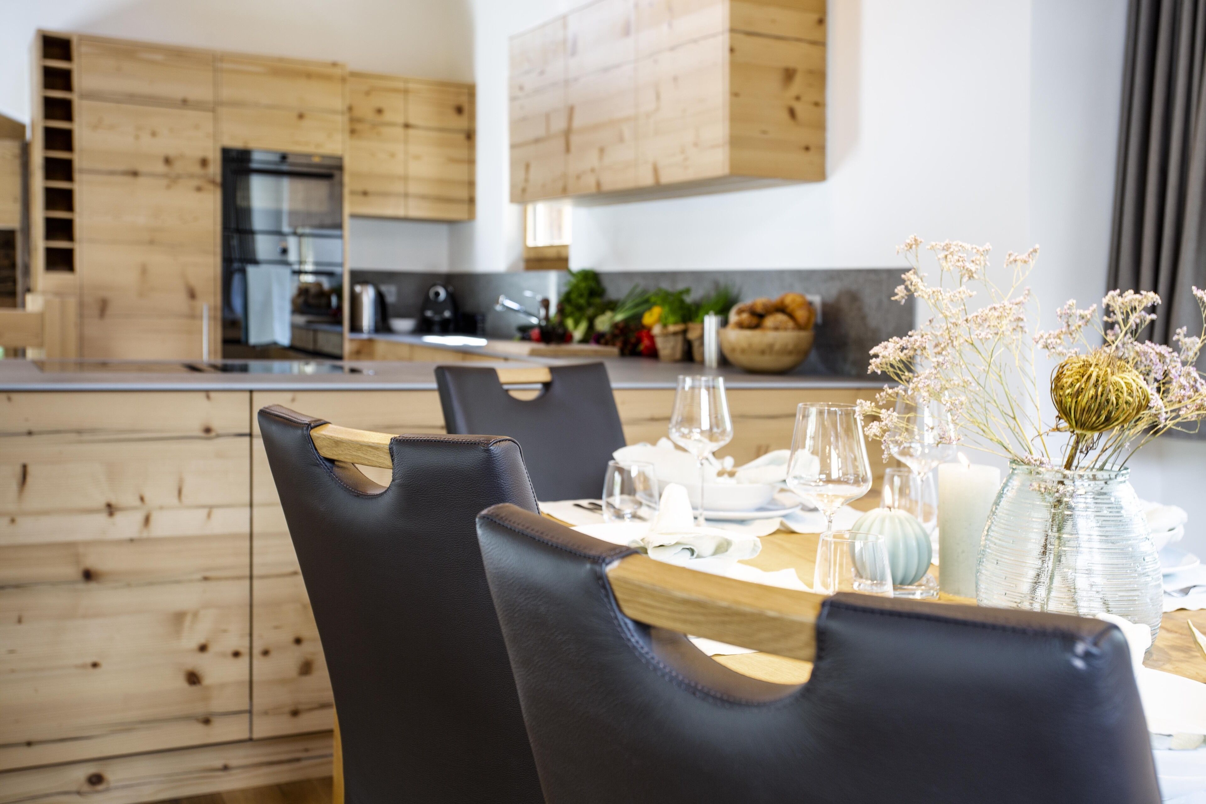 A modern kitchen with wooden cabinets, a dining area with a wooden table and chairs, and a vase with dried flowers on the table.