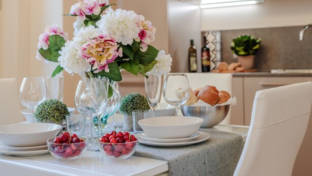 A dining table set with white plates, clear glass bowls with cherries, a vase with flowers, and wine glasses.