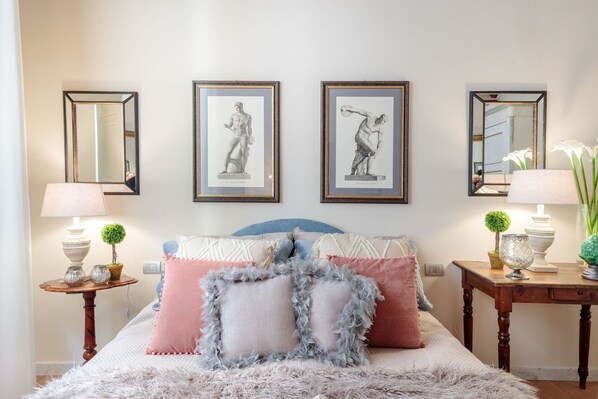 A bedroom with a bed, two framed pictures, two lamps, a small table, and a vase with flowers.