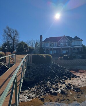 A large, two-story house with a stone facade and a red roof, situated next to a rocky shoreline. - Amazing Mountain, Lake and Island Views Private Beach,Fire Pit, Hot Tub, Kayaks  (Six Mile)