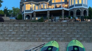 A large house with a stone facade and multiple windows. In front of the house, there is a stone wall and two kayaks with paddles. - Amazing Mountain, Lake and Island Views Private Beach,Fire Pit, Hot Tub, Kayaks  (Six Mile)