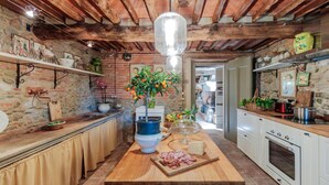 Une cuisine rustique aux murs en briques apparentes, avec des poutres en bois et un îlot central agrémenté d’un vase de fruits d’agrumes et d’une planche à fromages.