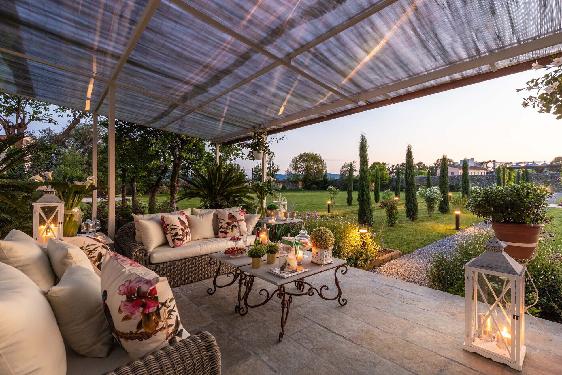 Un patio exterior con un área de estar techada, una mesa con velas y vistas a un jardín ajardinado.