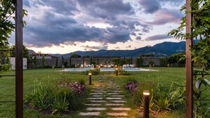 Un sentier de jardin mène à un espace piscine agrémenté d’une pergola et de sièges, le tout entouré d’une végétation luxuriante et avec des montagnes en toile de fond.