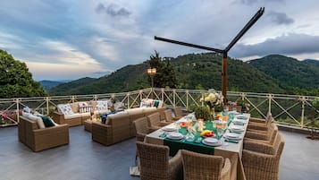 An outdoor dining area with wicker furniture and a table set for a meal, overlooking a mountainous landscape.