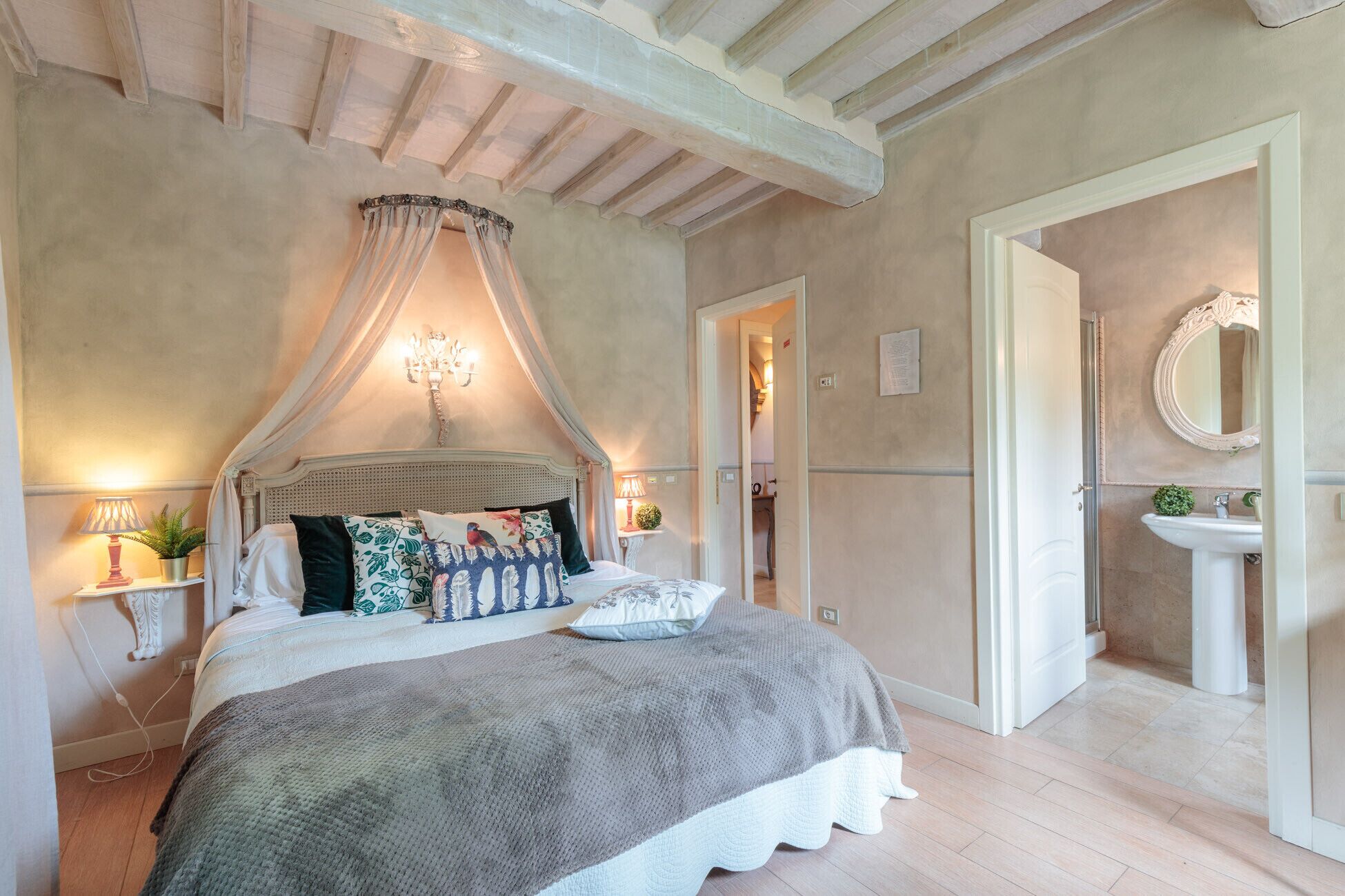 A bedroom with a canopy bed, bedside lamps, a bedside plant, and a view into the bathroom.