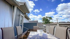A patio with a glass-top table and chairs, a building with a white exterior, and a clear blue sky with clouds.