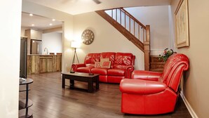 A living room with a red leather sofa, a coffee table, a clock on the wall, a ceiling fan, and a staircase leading to an upper level.