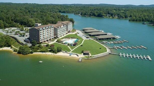 Vista aérea de un hotel con un edificio grande, un puerto deportivo con barcos y un lago rodeado de bosque.