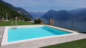 A rectangular pool with clear blue water, surrounded by a stone deck and a metal railing. In the background, there are mountains and a body of water.