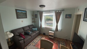 Ein Wohnzimmer mit einem braunen Sofa, gemusterten Kissen, Holzboden, einem Fenster mit Vorhängen und einem Wandbild.