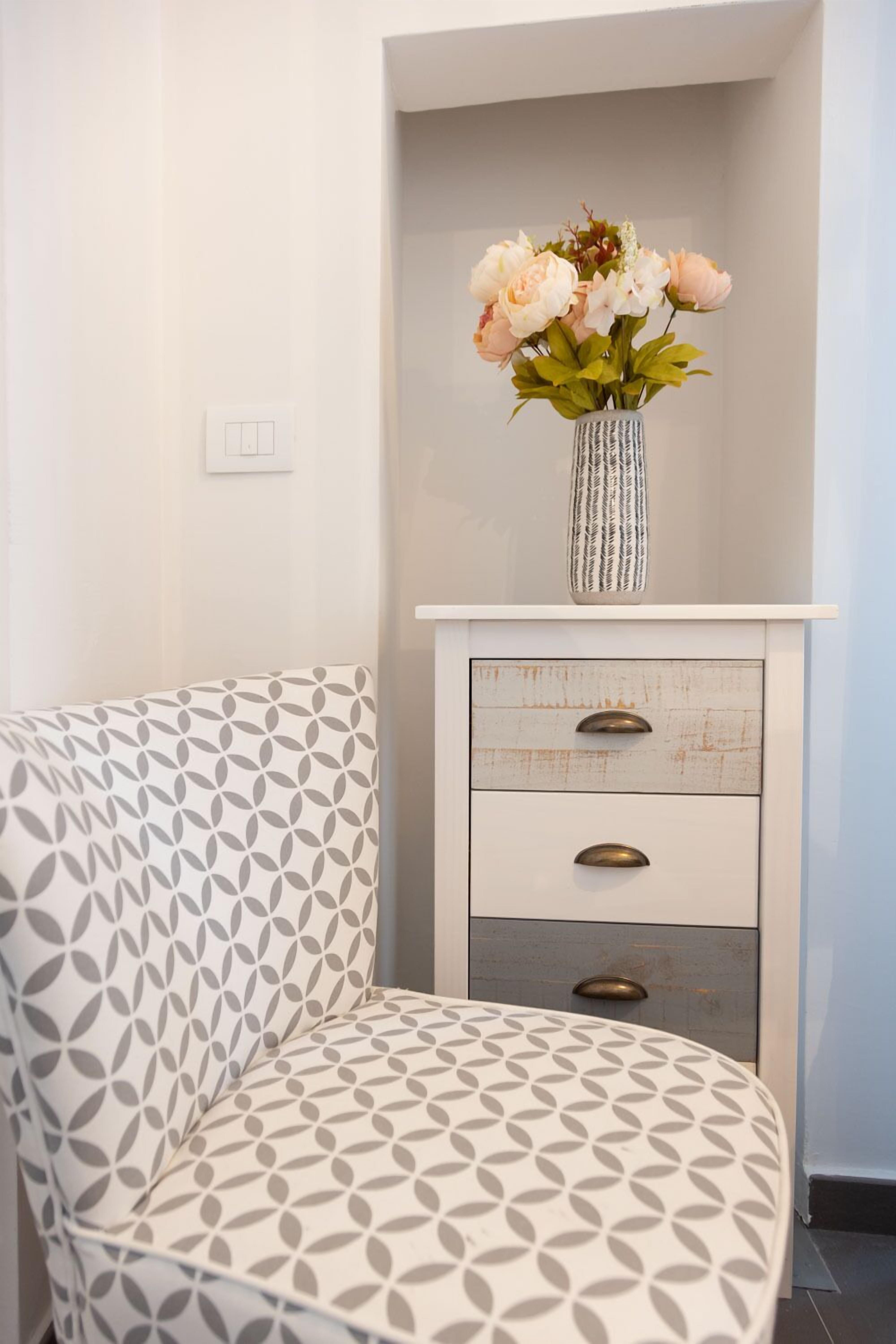 A white chair with a leaf pattern, a white dresser with three drawers, and a vase with flowers on top.