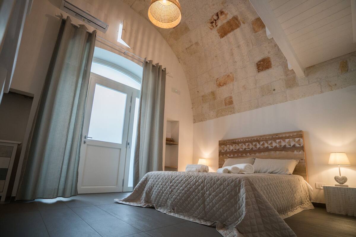 Ein Schlafzimmer mit einem großen Bett, einem hölzernen Kopfende, einem Nachttisch mit einer Lampe und Blick ins Freie durch ein Fenster.