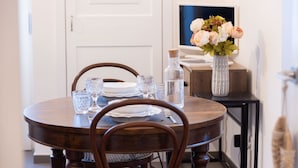 Un comedor con una mesa redonda de madera preparada para dos personas, con vasos y platos transparentes. Un jarrón con flores está sobre la mesa, y hay una botella de agua a un lado.