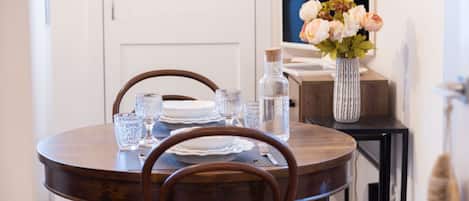 A dining area with a round wooden table set for two, equipped with clear glassware and plates. A vase with flowers is on the table, and a bottle of water is on the side.