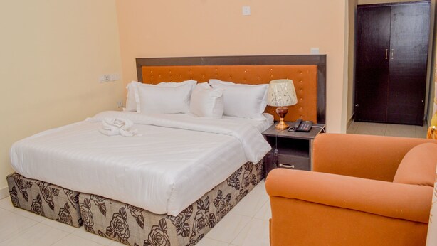 A hotel room with a large bed, a bedside table with a lamp, and an orange armchair. - Kedros Precious Suites (Ilorin)