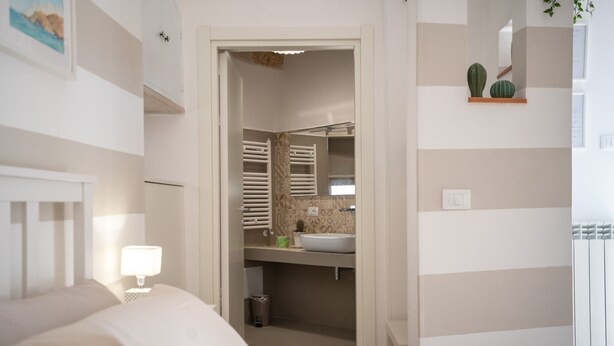 A hotel room with a bed, a bedside table with a lamp, a bathroom with a sink and towel racks, and a small shelf with decorative items. - Mest Michè by Polignando (Polignano a Mare)