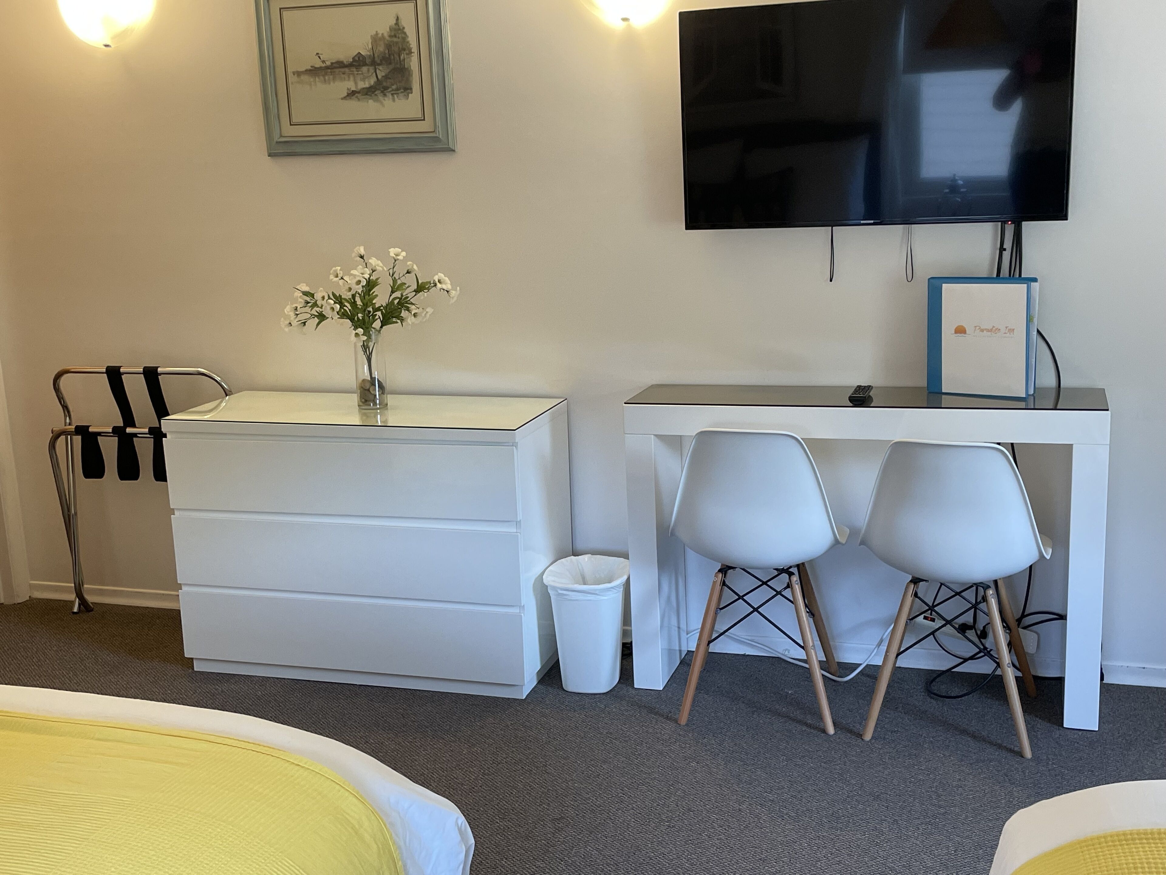 Une chambre d’hôtel comprenant un lit, un bureau avec deux chaises, une télévision, une commode, une poubelle et un tableau accroché au mur.