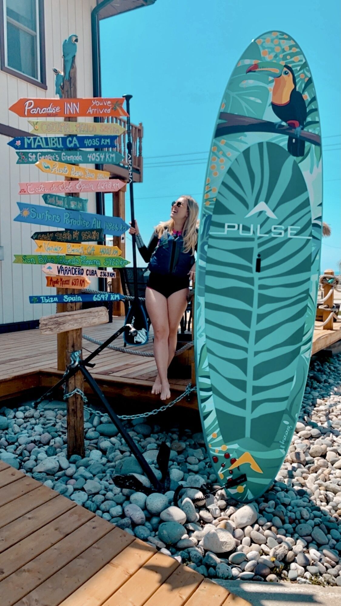 Una mujer de pie junto a una tabla de surf con un diseño de hojas tropicales, sosteniendo un cartel con varios nombres de destinos y distancias.