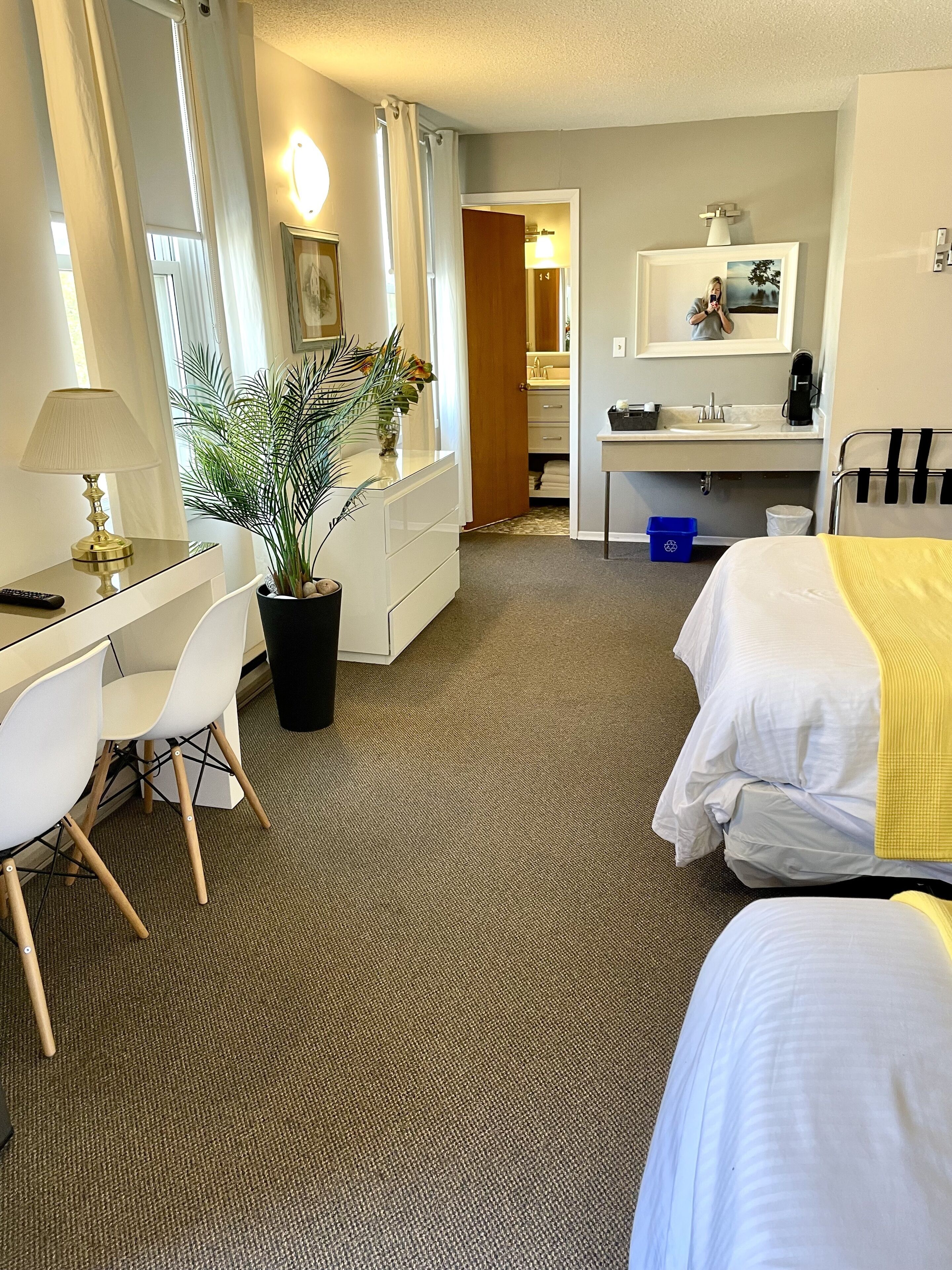 Habitación de hotel con escritorio, dos camas, una planta en maceta, un espejo y baño visible a través de una puerta abierta.