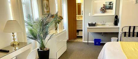 Habitación de hotel con escritorio, dos camas, una planta en maceta, un espejo y baño visible a través de una puerta abierta.