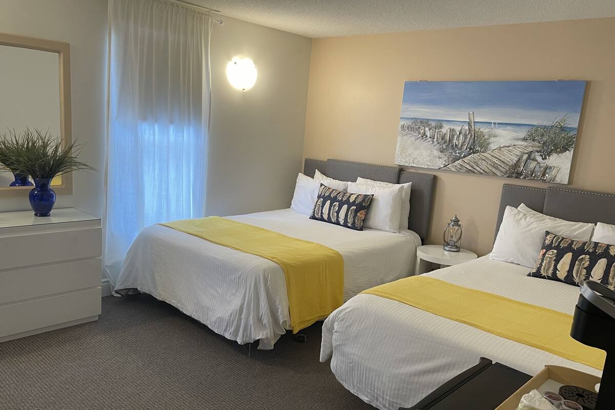 A hotel room with two beds, a desk, a lamp, a painting, and a potted plant.
