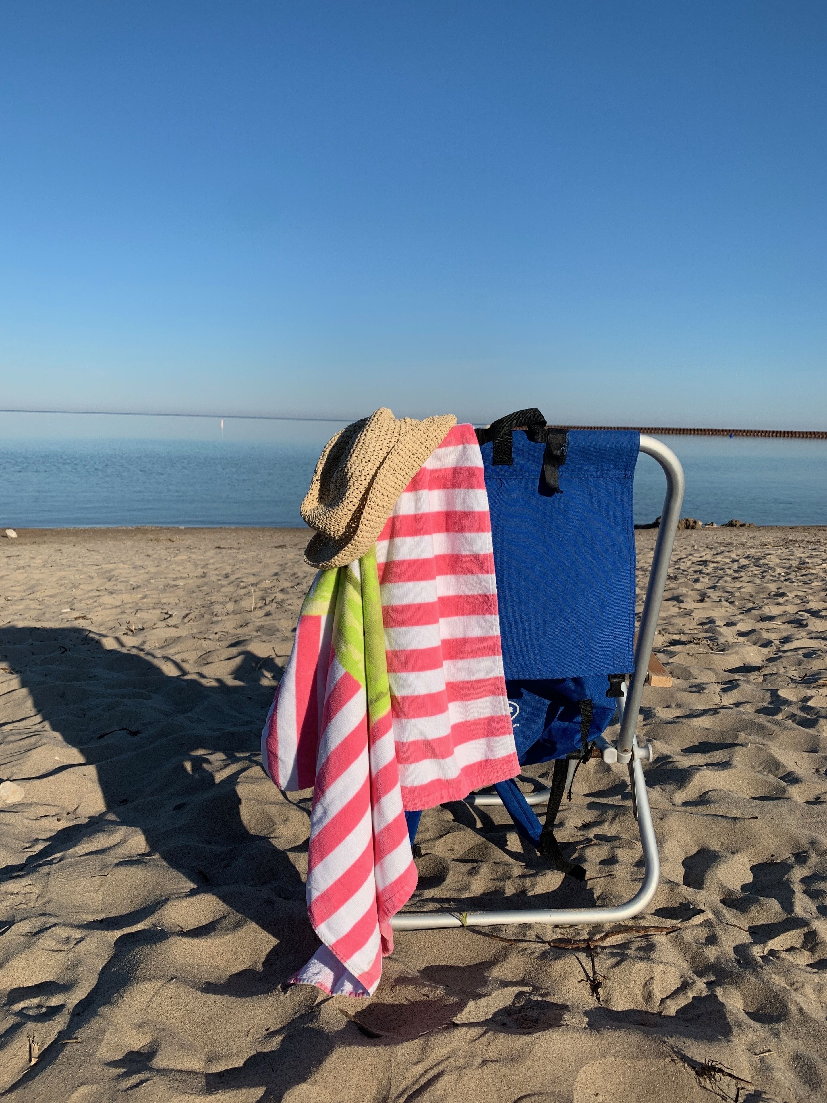 A beach chair with a straw hat and striped towels on a sandy beach.