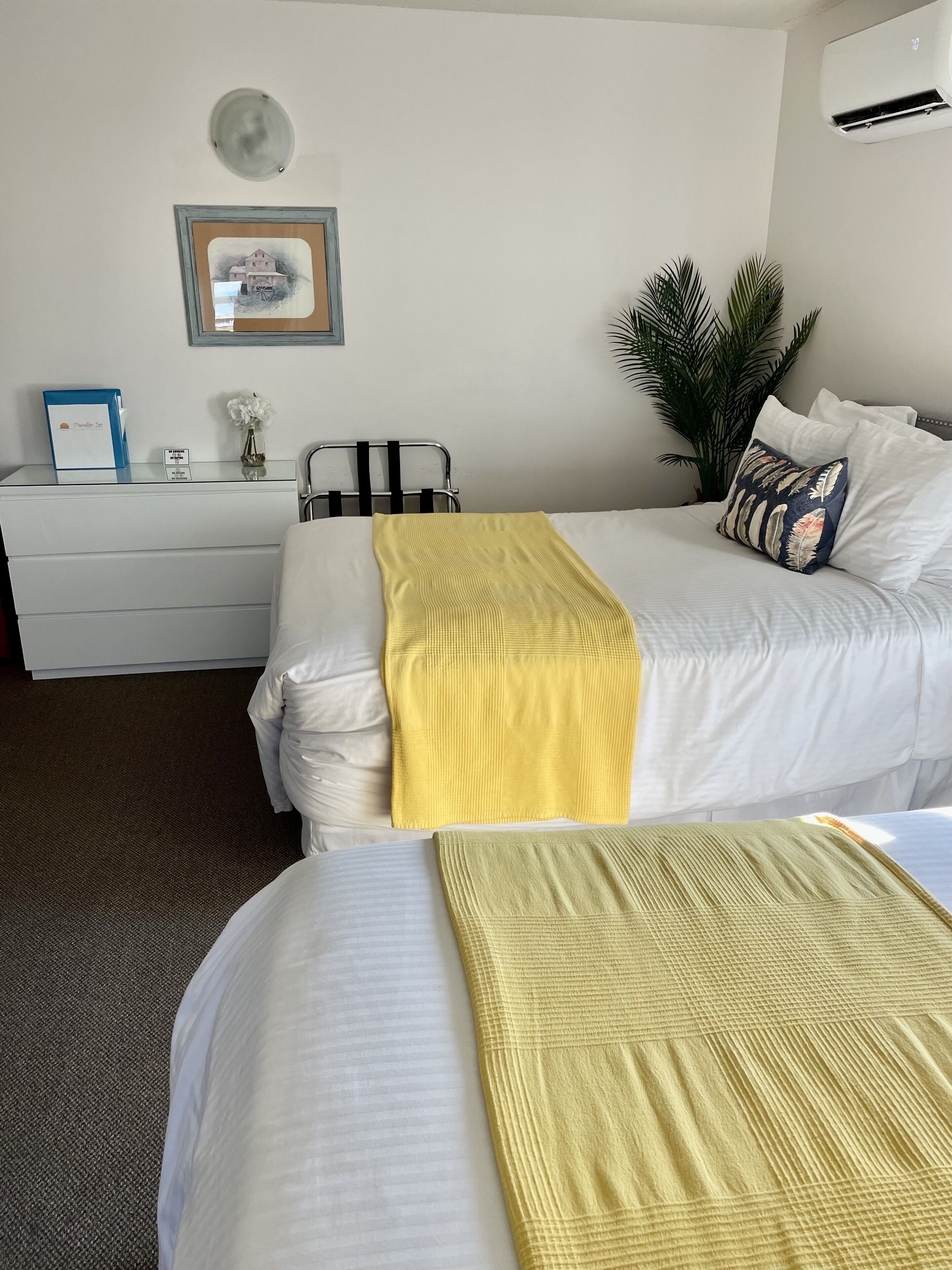 A hotel room with a bed, a bedside table, a lamp, a framed picture, and a potted plant.