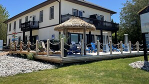A two-story building with a balcony, a thatched umbrella, and a wooden deck with chairs. - Paradise Inn On The Beach (Saugeen Shores)