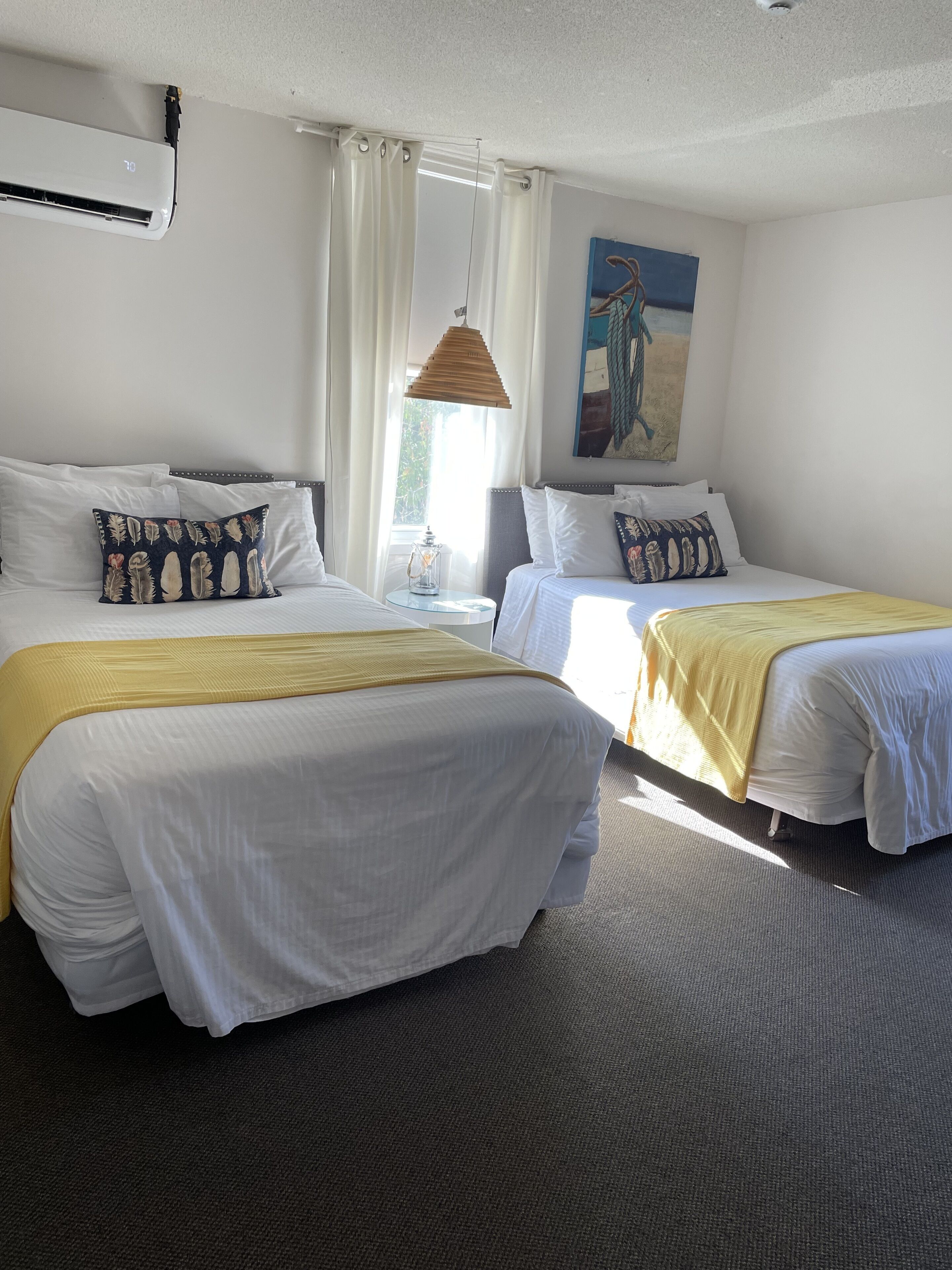 Habitación de hotel con dos camas, una ventana con cortinas, un cuadro en la pared y un equipo de aire acondicionado.