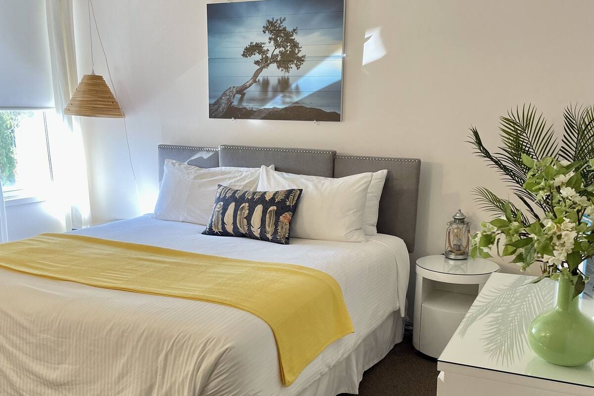 A neatly made bed with a yellow throw blanket, a grey headboard, and a wall-mounted picture of a tree.