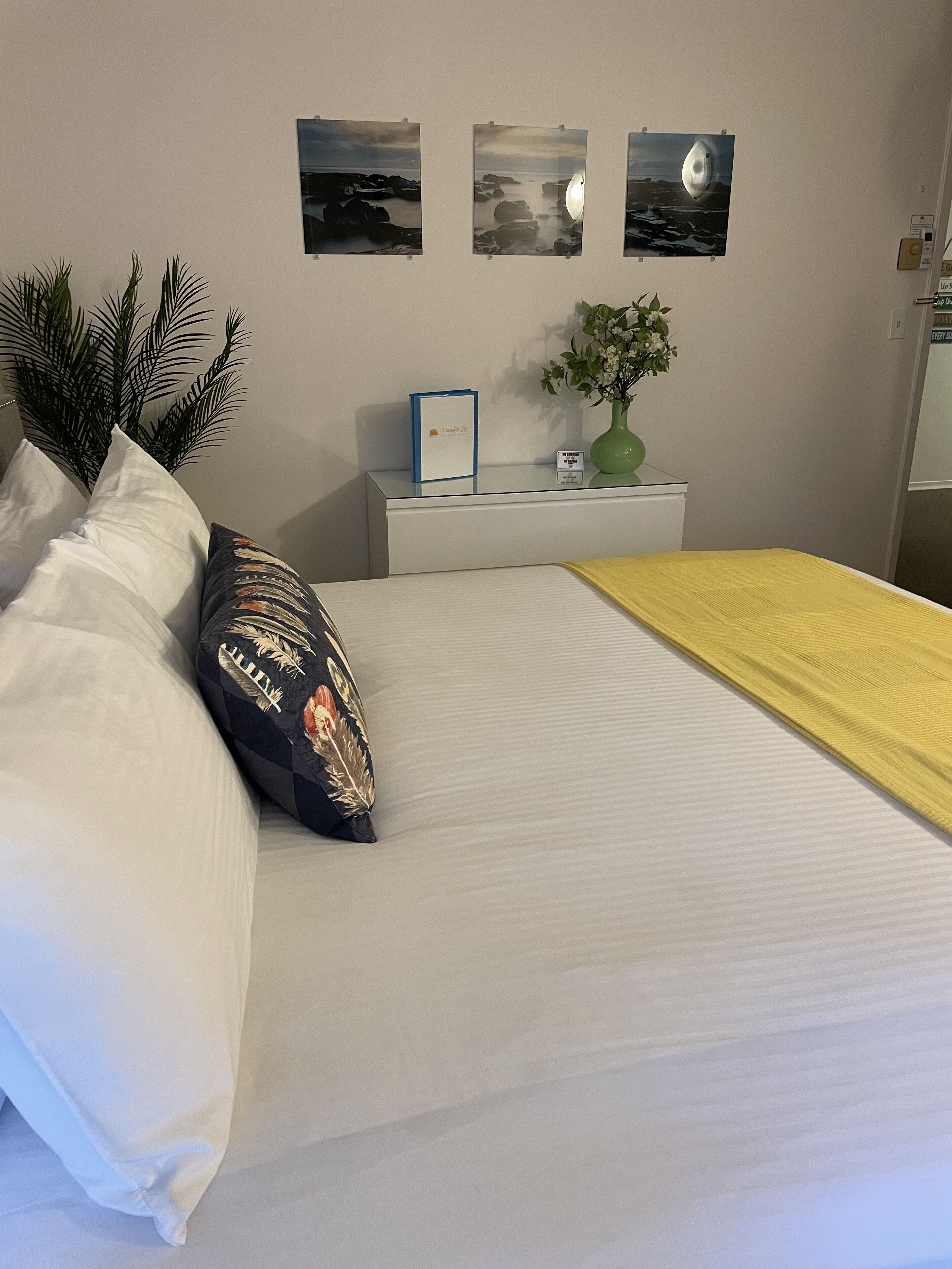 A neatly made bed with white and yellow bedding, a black and gold pillow, and a green plant on a bedside table. There are three framed pictures on the wall above the bed.