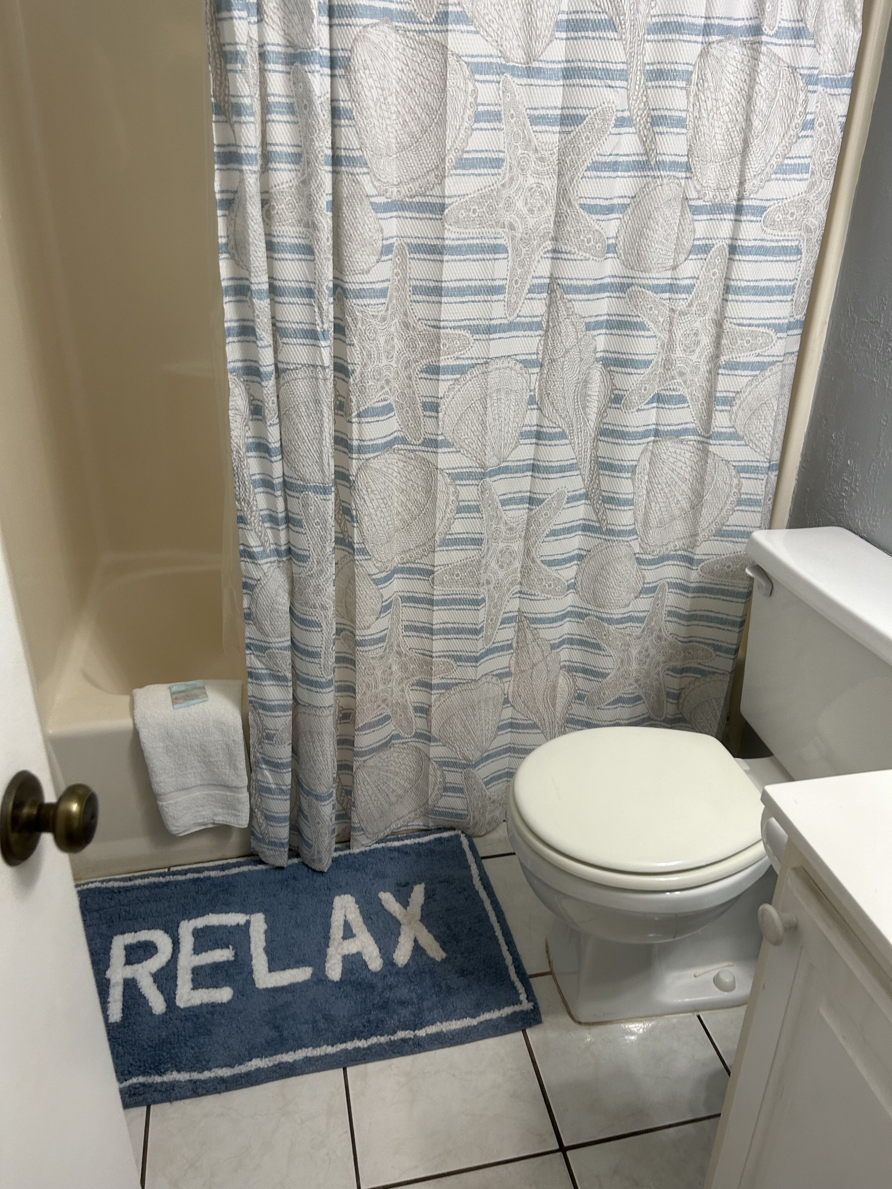 A bathroom with a toilet, a shower curtain with a striped pattern, and a blue mat with the word "RELAX" on it.