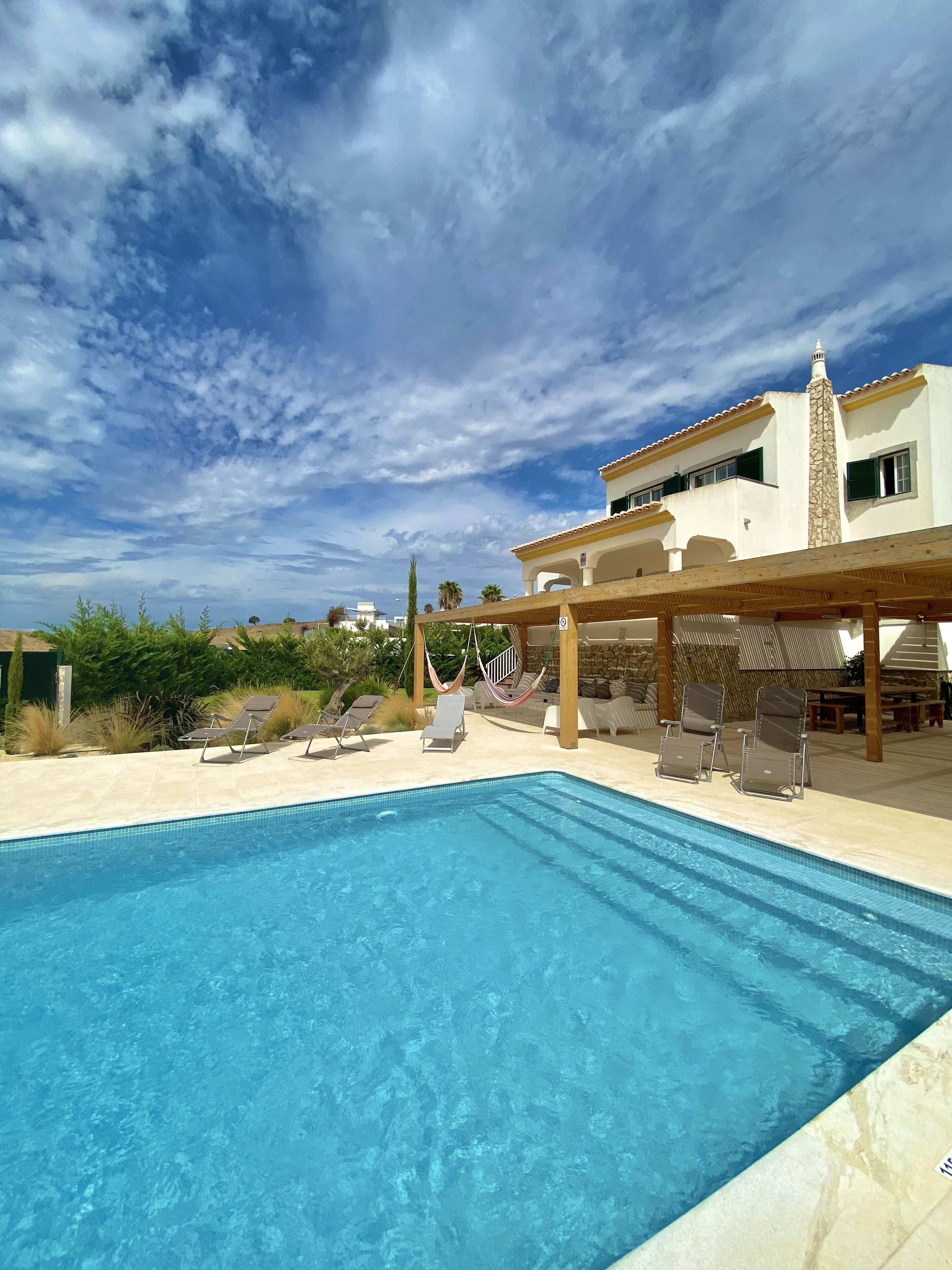 A swimming pool with lounge chairs and a pergola, surrounded by a building with a stone facade and a clear blue sky.