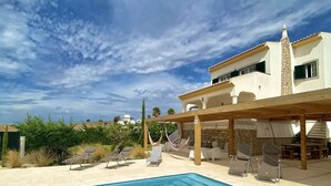 Ein Swimmingpool mit Liegestühlen und einer Pergola, umgeben von einem Gebäude mit Steinverkleidung und blauem Himmel.